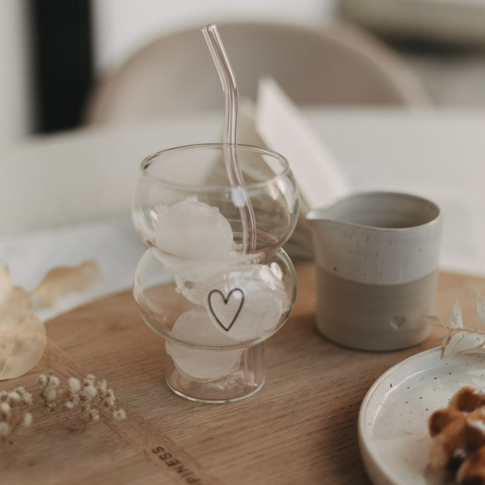 Eulenschnitt Bubble Glas mit schwarzem Herz-Print, gefüllt mit Iced Coffee und Eiswürfeln. Daneben ein Keramikkännchen und ein Teller mit belgischer Waffel auf einem runden Holzbrett.