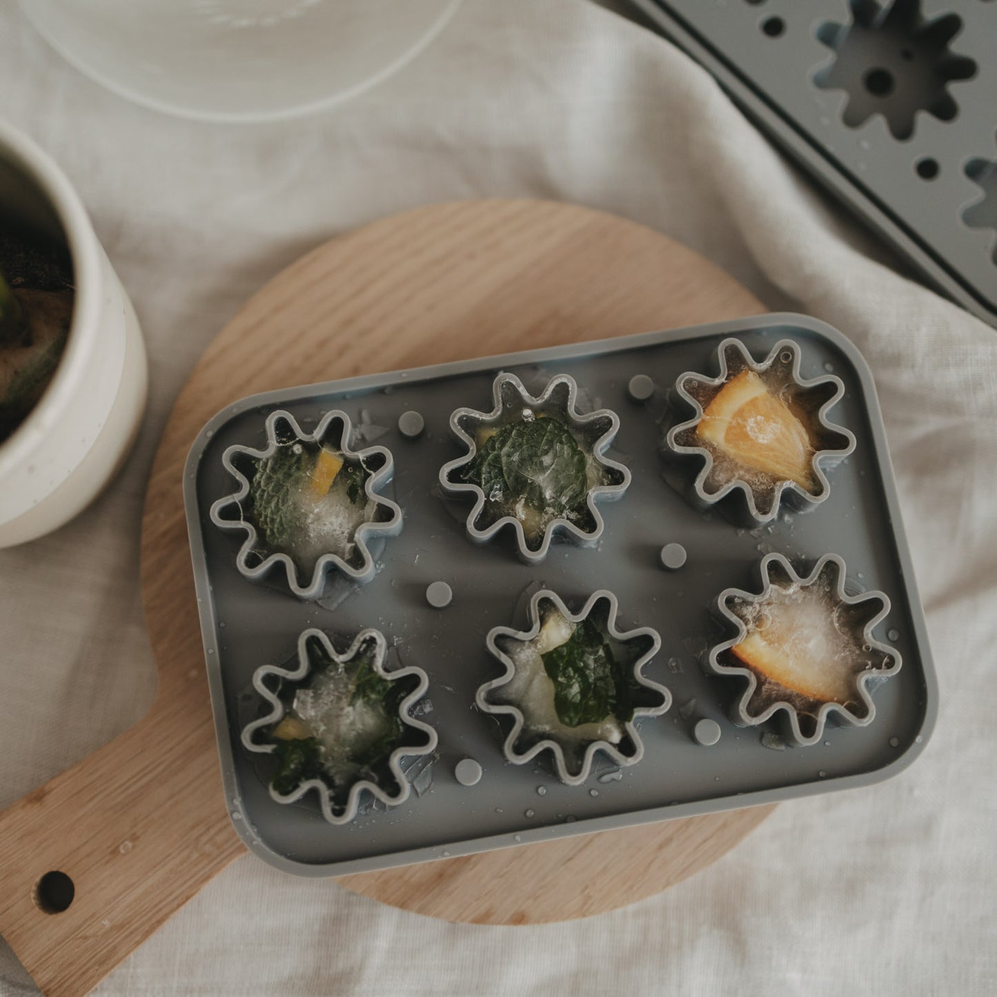 Fertige Eiswürfel mit Orangenscheiben oder Minze liegen in der grauen Blütenform aus flexiblem Silikon, dekoriert auf einem runden Holzbrett.
