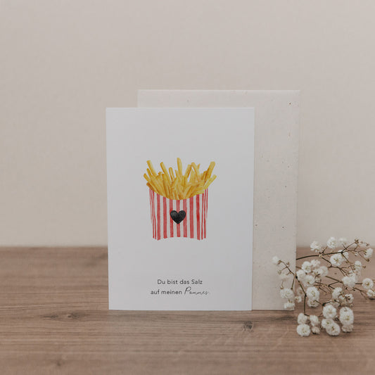 Klappkarte mit Illustration von Pommes in rot-weiß gestreifter Verpackung und Text 'Du bist das Salz auf meinen Pommes' auf Holztisch mit Umschlag und weißen Blüten