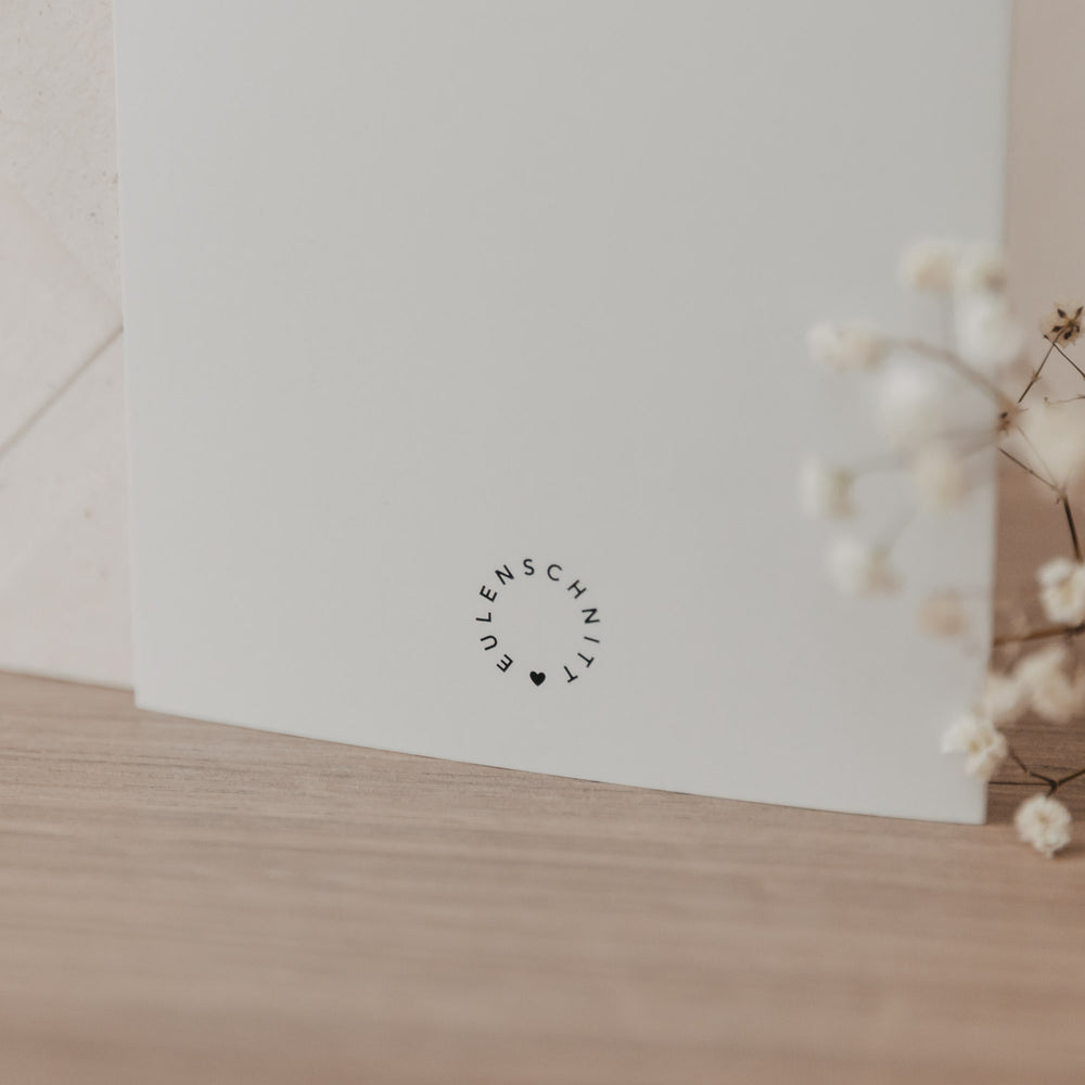 Klappkarte von hinten mit kleinen runden Eulenschnitt Logo mit minimalistischem Design auf Holzuntergrund, dekoriert mit getrockneten Blumen