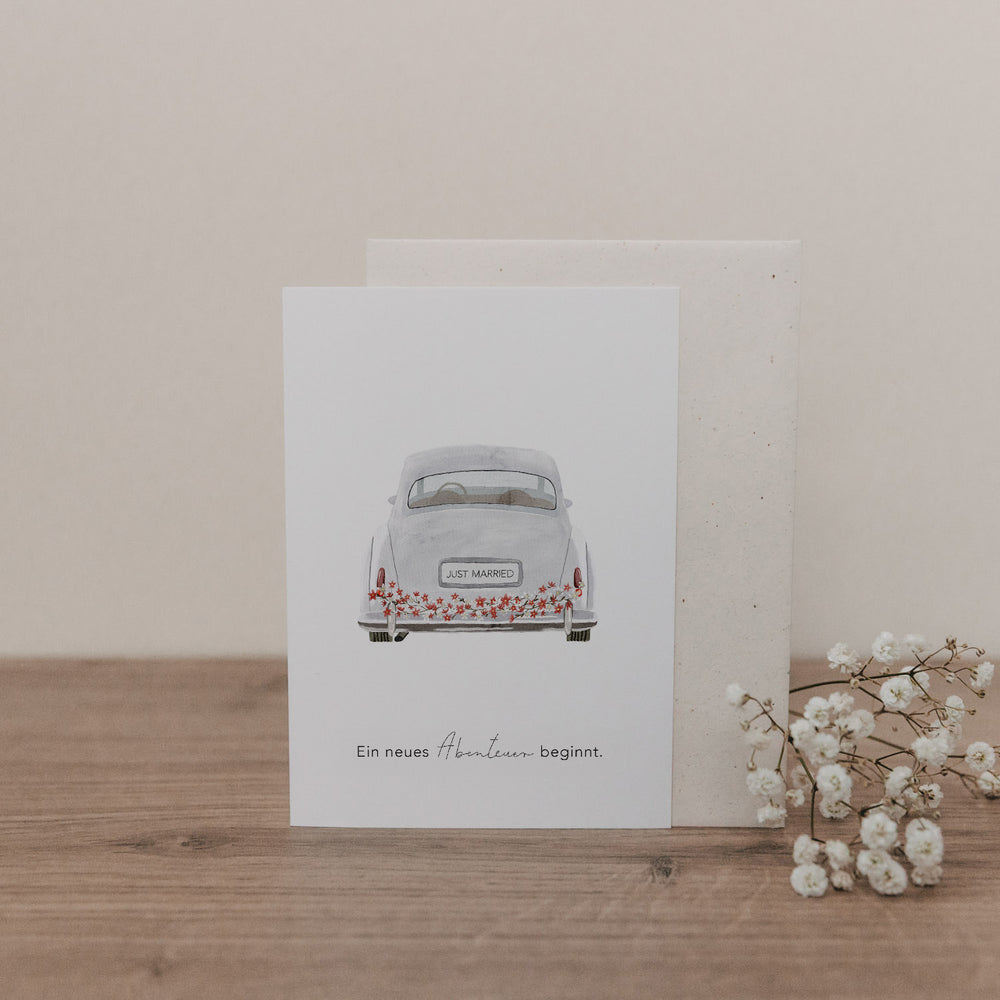 Klappkarte mit Illustration eines weißen Hochzeitsautos von hinten, Nummernschild 'Just Married', Text 'Ein neues Abenteuer beginnt' auf Holzuntergrund mit Schleierkraut
