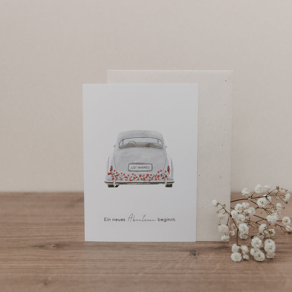 Klappkarte mit Illustration eines weißen Hochzeitsautos von hinten, Nummernschild 'Just Married', Text 'Ein neues Abenteuer beginnt' auf Holzuntergrund mit Schleierkraut