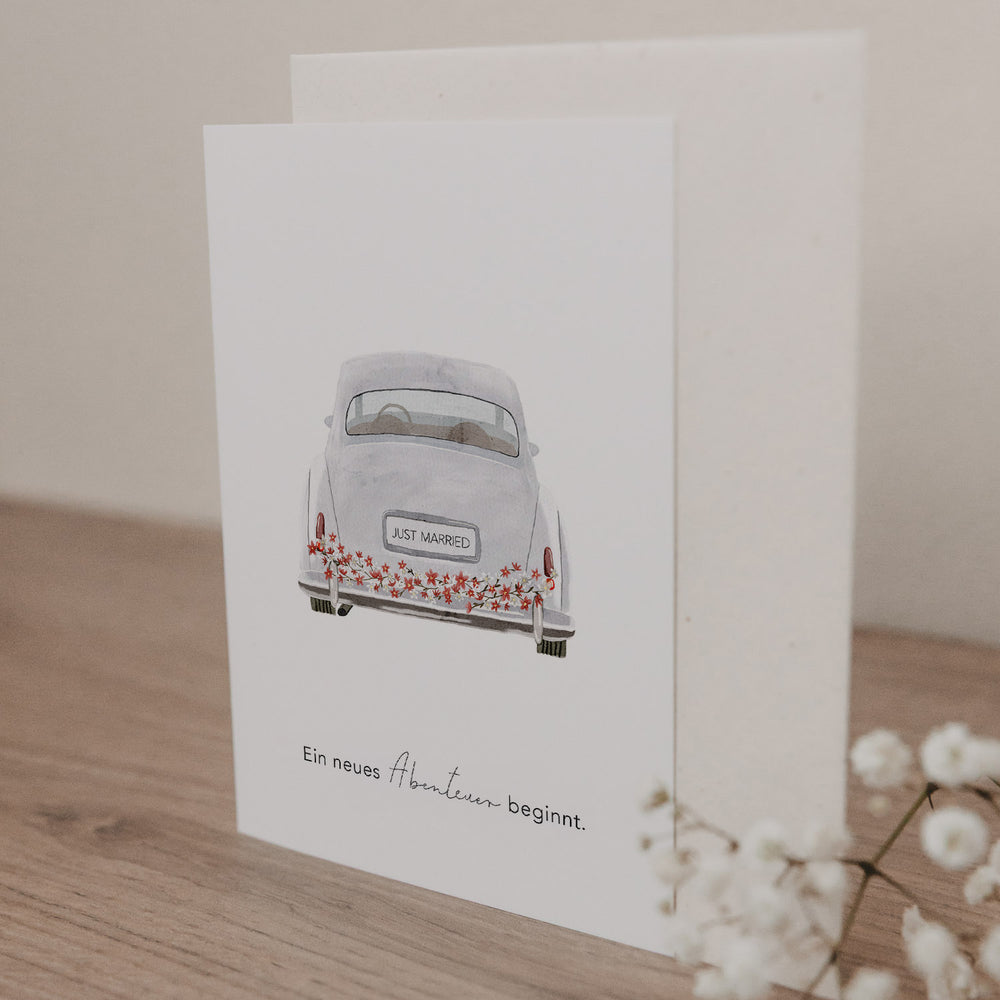 Klappkarte mit Illustration eines Hochzeitsautos und dem Schriftzug 'JUST MARRIED', darunter Text 'Ein neues Abenteuer beginnt'