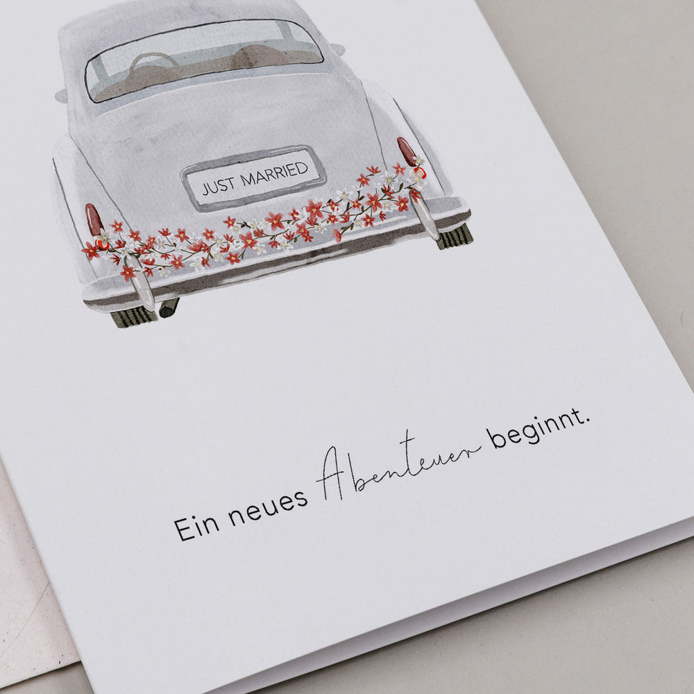 Klappkarte mit Aquarellzeichnung eines Hochzeitsautos mit 'JUST MARRIED'-Kennzeichen und Blumenschmuck, darunter der Text 'Ein neues Abenteuer beginnt.'