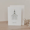 Klappkarte mit handgezeichneter Kirche und Schrift „Alles Liebe“, stehend mit Umschlag auf Holzoberfläche neben Trockenblumen.