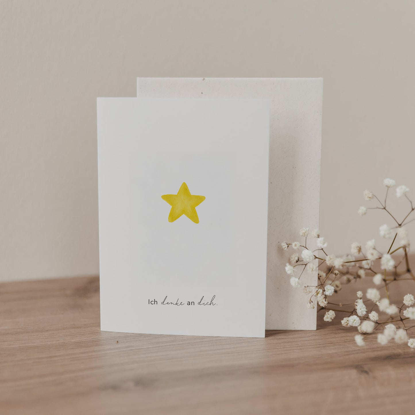 Klappkarte mit gelbem Stern und Schrift „Ich denke an dich“, stehend mit Umschlag auf Holzoberfläche dekoriert mit Trockenblumen.