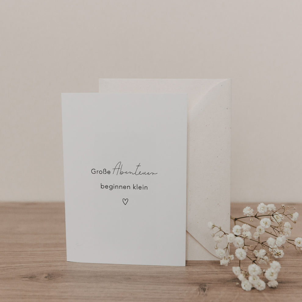 Klappkarte „Große Abenteuer beginnen klein“ in Weiß mit Herzmotiv und passendem Umschlag, dekoriert mit zarten Blüten.