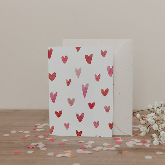Klappkarte mit rosanen und roten Aquarellherzen auf weißem Hintergrund, inklusive Umschlag, dekoriert mit Konfetti und Blumen.