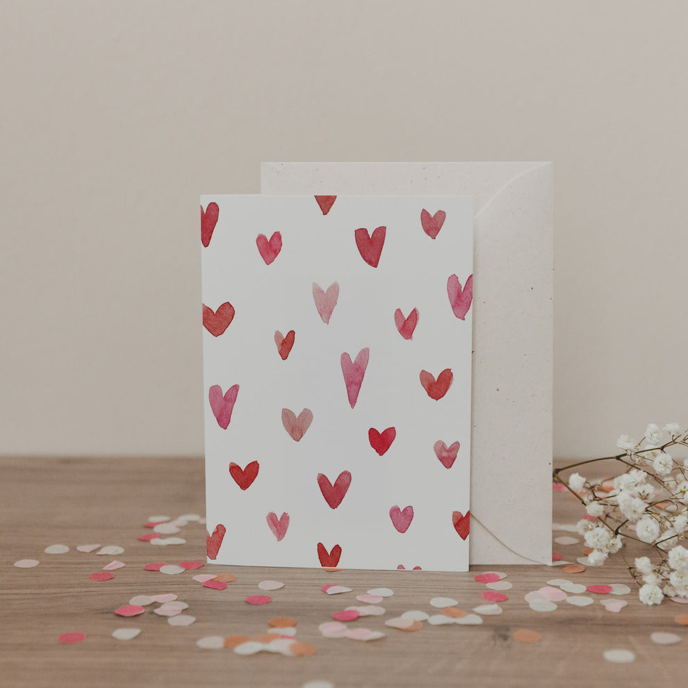 Klappkarte mit rosanen und roten Aquarellherzen auf weißem Hintergrund, inklusive Umschlag, dekoriert mit Konfetti und Blumen.