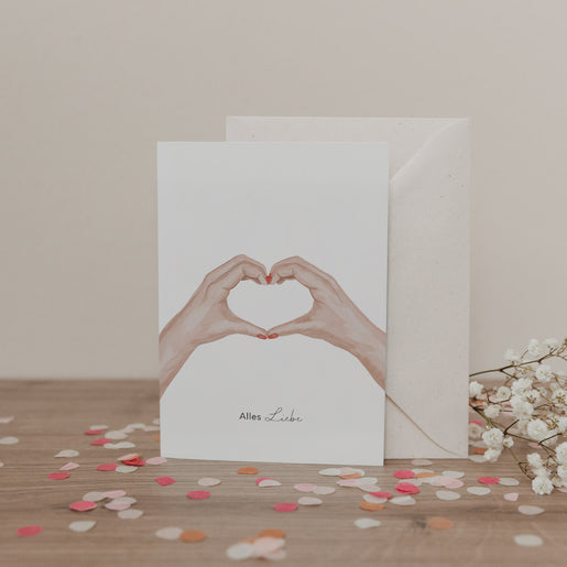 Klappkarte mit zwei Händen in Herzform und dem Schriftzug „Alles Liebe“, vor Umschlag und Blumen, auf hellem Untergrund.