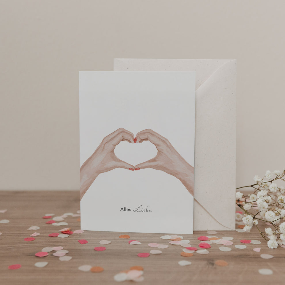 Klappkarte mit zwei Händen in Herzform und dem Schriftzug „Alles Liebe“, vor Umschlag und Blumen, auf hellem Untergrund.
