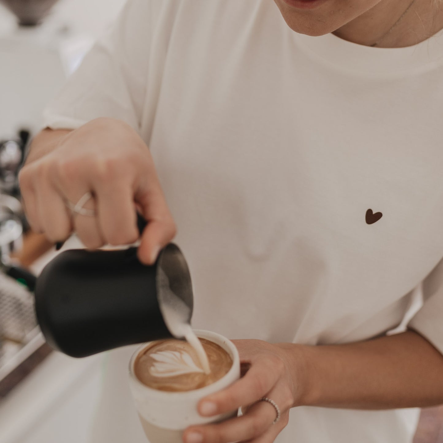 Nahaufnahme: Frau gießt Latte Art in Kaffeetasse, Herzmotiv auf T-Shirt sichtbar.