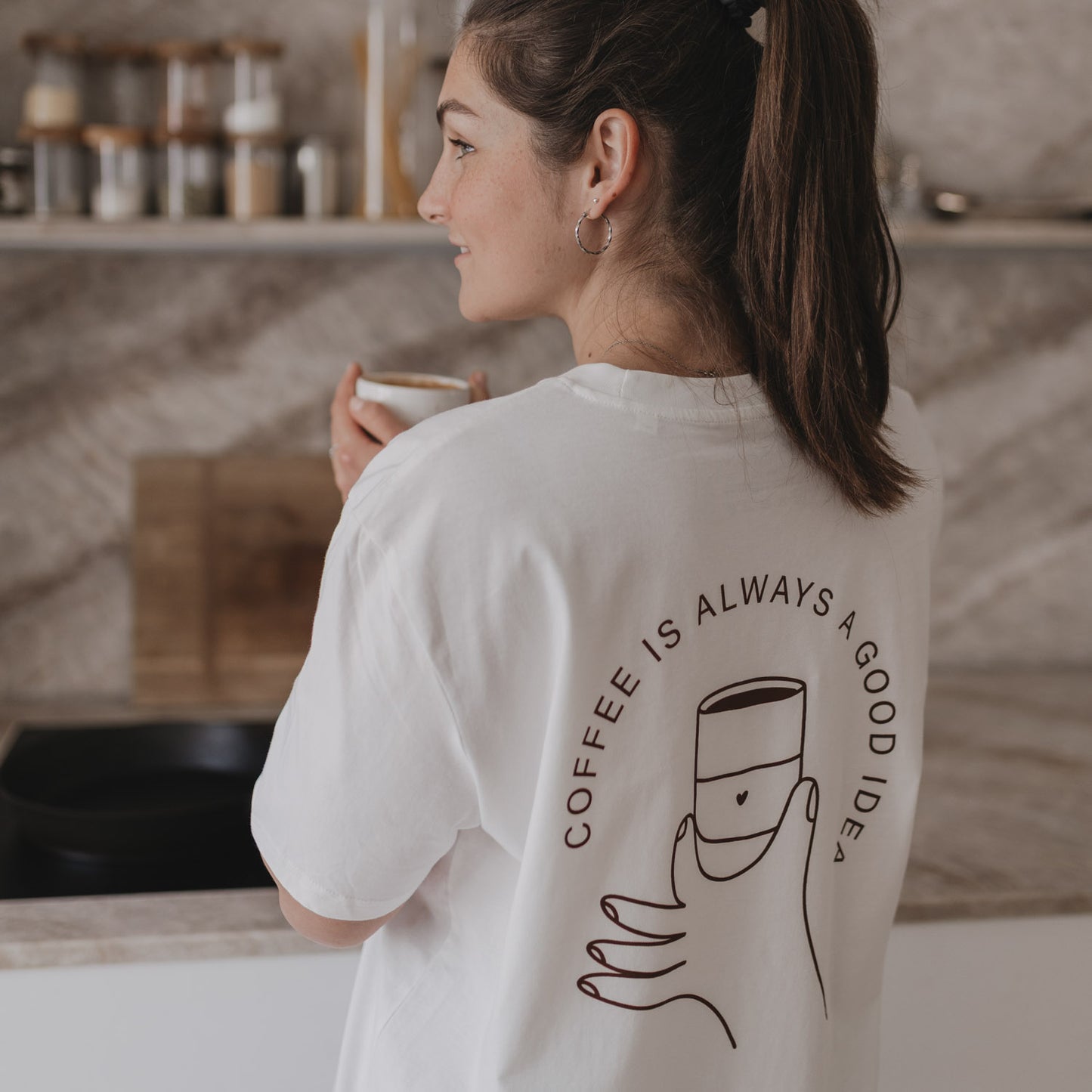 Seitliche Ansicht: Frau mit Tasse in der Hand, Rückseite des Shirts mit Kaffeetassenmotiv gut sichtbar.