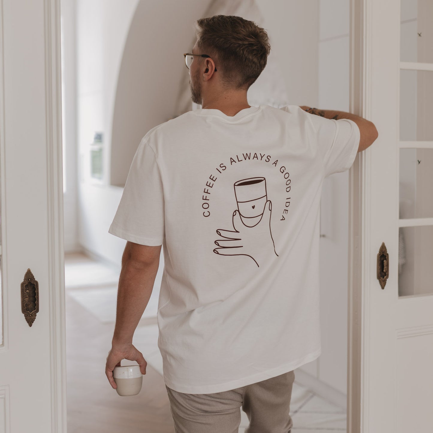 Rückansicht eines Mannes mit Coffee T-Shirt beim Kaffeetrinken in heller Küche mit Glastür