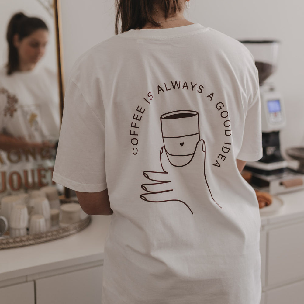 Person mit weißem T-Shirt zeigt Rückseite mit Illustration einer Hand, die eine Tasse hält und dem Schriftzug.