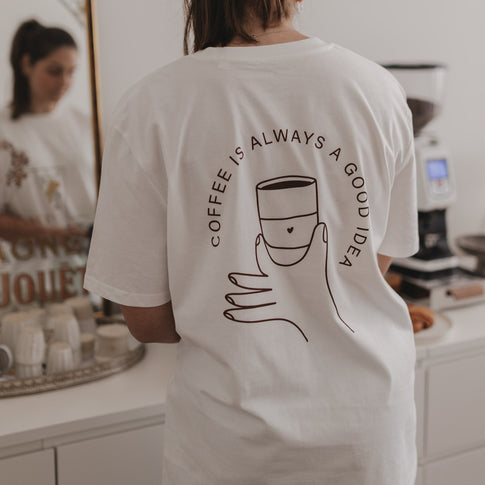 Person mit weißem T-Shirt zeigt Rückseite mit Illustration einer Hand, die eine Tasse hält und dem Schriftzug.