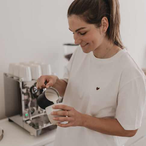 Frau trägt weißes T-Shirt mit Herz-Print auf der Brust und bereitet Kaffee mit Siebträgermaschine zu.