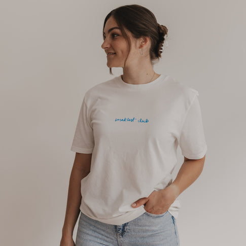 Frau trägt weißes Frühstücks-Shirt mit blauem Schriftzug, blickt seitlich in die Kamera.