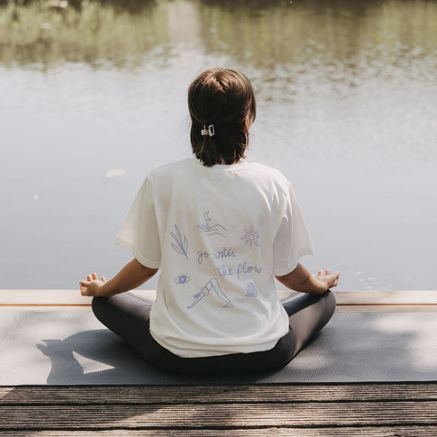 Frau meditiert am See auf Yogamatte, trägt weißes T-Shirt „go with the flow“ mit Rückendruck