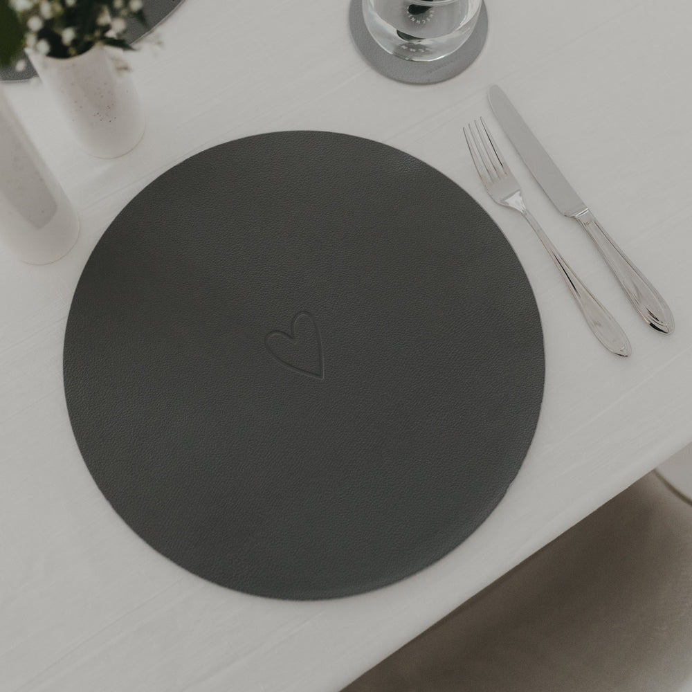Dunkelgraues rundes Platzset mit eingeprägtem Herzmotiv, stilvoll gedeckt mit Besteck auf weißer Tischdecke.