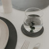 Glas auf dunkelgrauem Untersetzer mit Prägung ‚Das Leben ist schön‘ auf hell gedecktem Tisch.