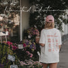 Frau im weißen „Coffee Lover“-T-Shirt und rosa Cap mit Iced Coffee vor einem Blumenladen.
