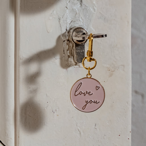 Ein Schlüsselanhänger in der Farbe rosa & gold mit der Aufschrift *love you* hängt an einem Schlüssel, der in einer schönen alten weissen Tür steckt.