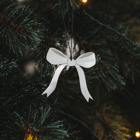 Anhänger in Schleifenform aus mattem Silber hängt an grünem Tannenzweig mit Lichterkette.