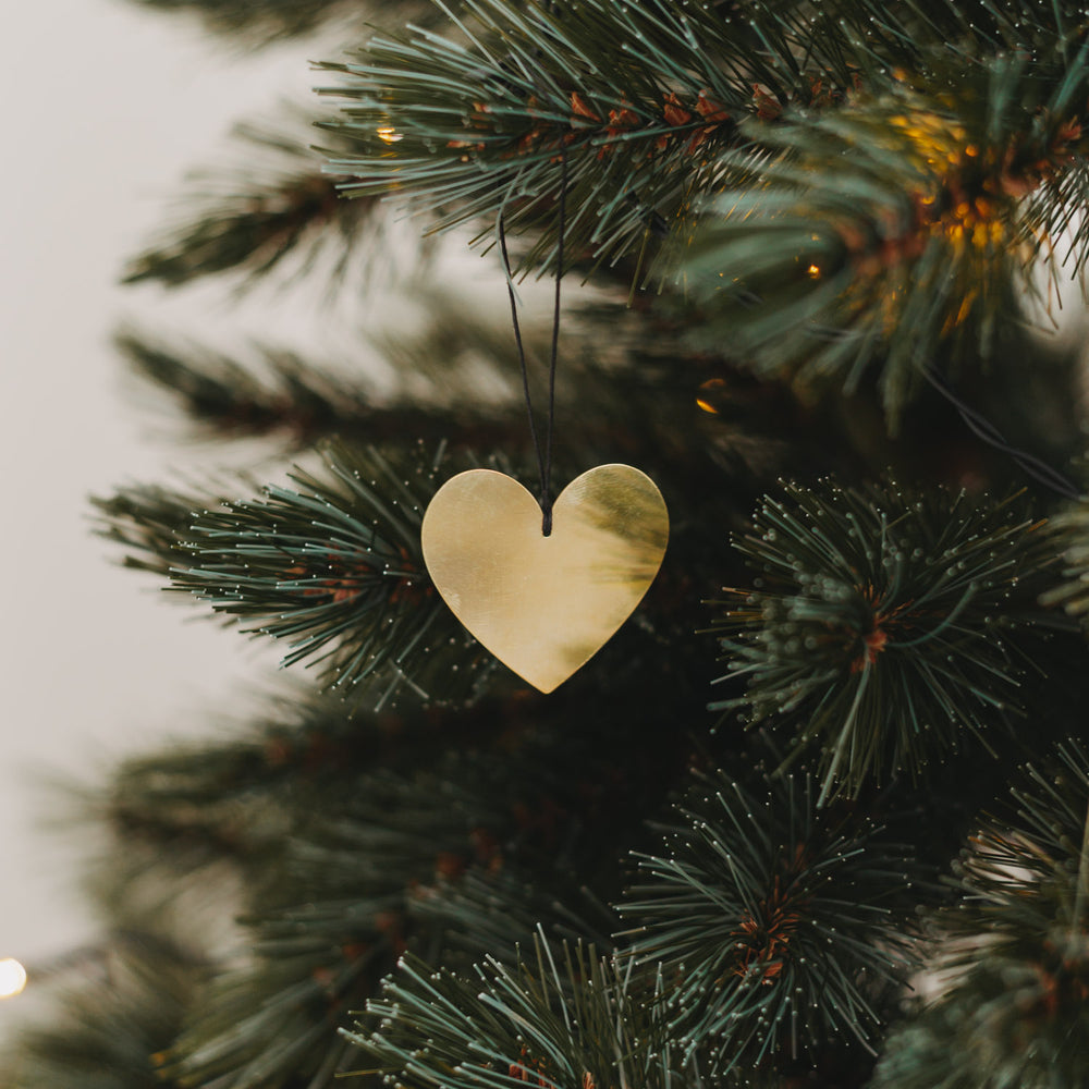 Goldenes Herz aus Metall am schwarzen Band, dekorativ am grünen Tannenbaum mit Lichterkette aufgehängt.