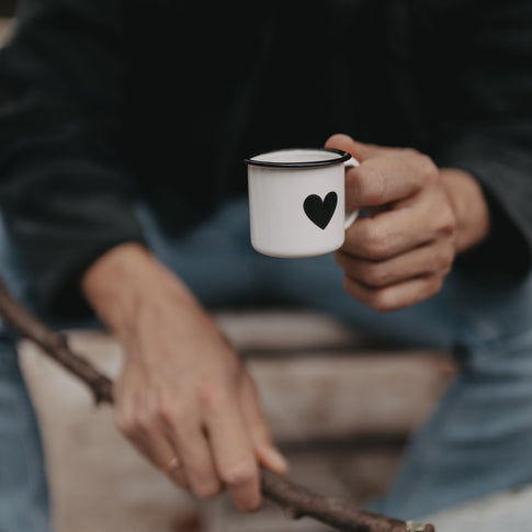 Hand hält Emaille Espressotasse Herz am Lagerfeuer – stilvolle kleine Tasse für Espresso und Outdoor-Genuss.