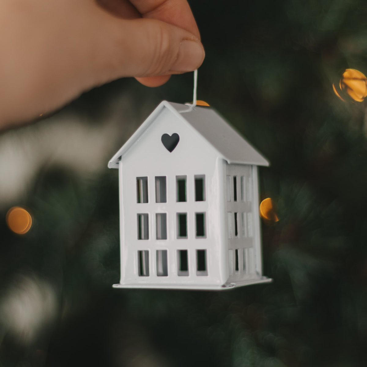 Weißer Hausanhänger aus Metall mit Herzdetail und weißer Kordel vor warmen Weihnachtslichtern