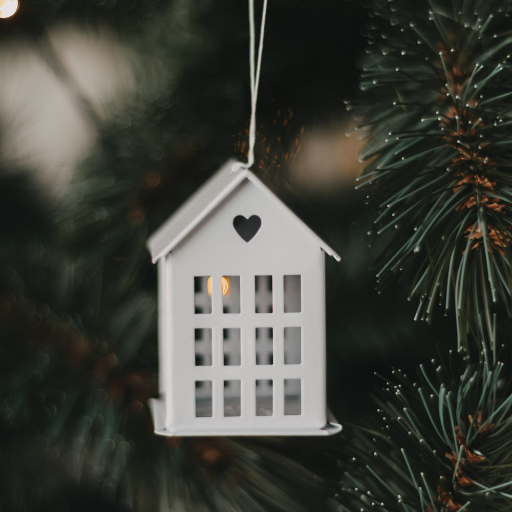 Weißer Metallanhänger in Hausform mit Herzdetail am festlich beleuchteten Weihnachtsbaum