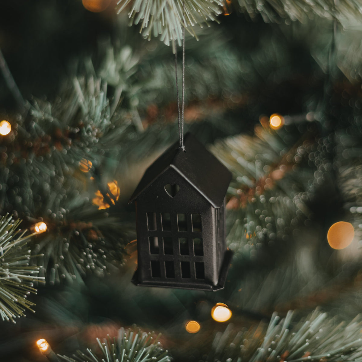 Schwarzer Metallanhänger in Hausform mit Herzdetail und Kordel am beleuchteten Weihnachtsbaum