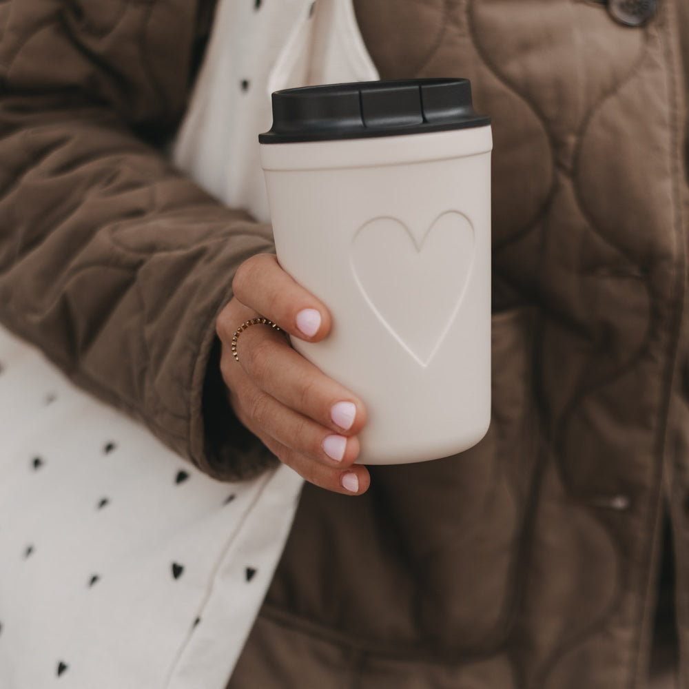 Frau mit cremefarbenem To-Go-Becher mit Herzmotiv und Stofftasche 