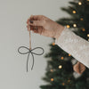 Metallanhänger Schleife in Anthrazit mit Juteschnur als Weihnachtsbaumschmuck vor einem beleuchteten Tannenbaum.