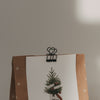 Nahaufnahme eines schwarzen Foldback-Clips mit Herzmotiv, der eine Weihnachtskarte an einer Geschenktüte befestigt.