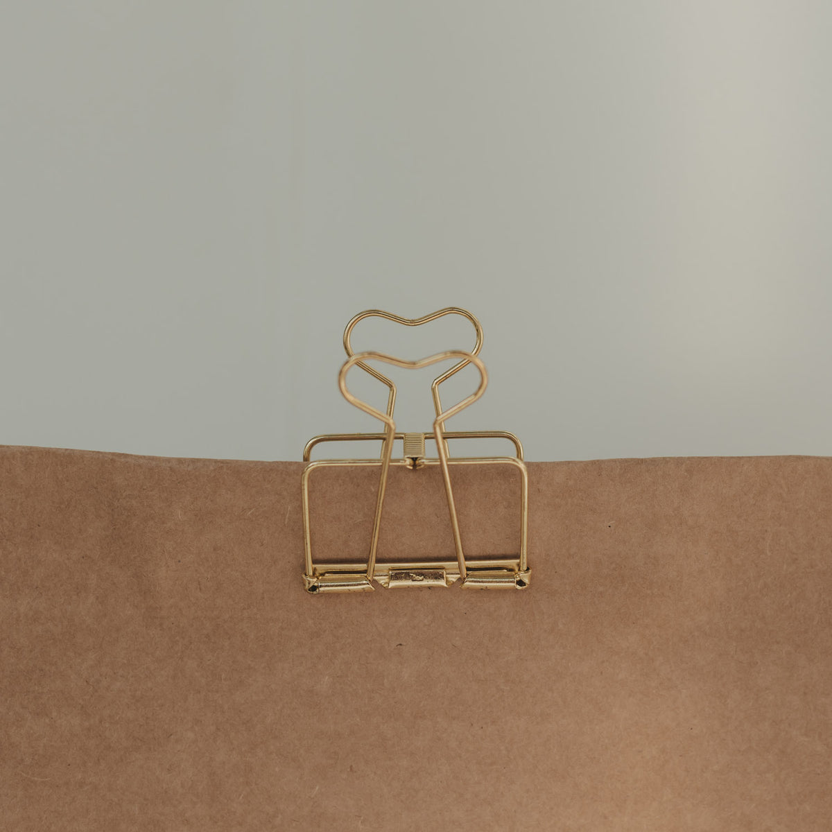 Große goldene Foldback-Klammer mit herzförmigen Griffen verschließt eine braune Geschenktüte aus Kraftpapier.