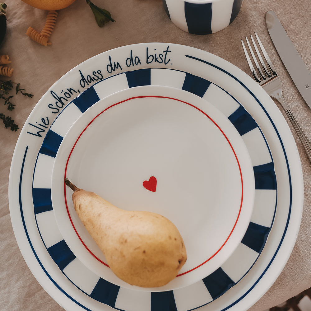 Gestapelte Teller mit blauem Streifenrand, Schriftzug „Wie schön, dass du da bist“ und roten Herz inszeniert mit Birne und Besteck auf natürlicher Leinentischdecke