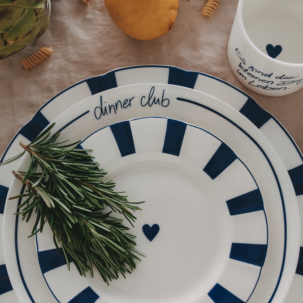 Gestapelte Teller mit blauem Streifenrand und 1 Teller mit „dinner club“ Schriftzug, dekoriert mit Rosmarinzweig und kombiniert mit passendem Becher auf Leinentischdecke