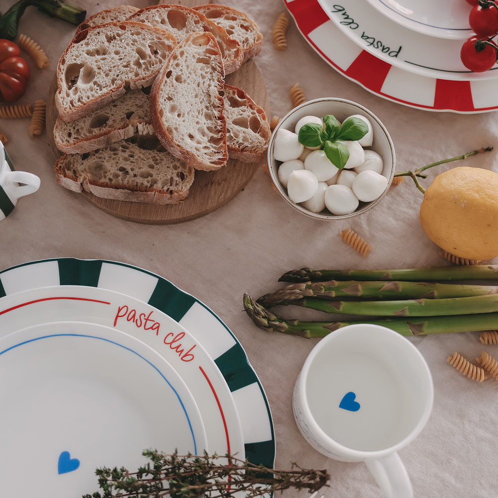 Stilvolles Mix & Match aus Porzellan mit buntem Streifenmuster, "Pasta Club"-Schrift und Herzmotiv, inszeniert mit frischen Zutaten wie Brot, Kräutern und Gemüse.