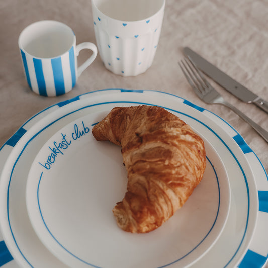 Weißer „breakfast club“-Teller mit blauem Rand und Croissant, dazu blau-weiß gestreifte Tasse, Teller, Becher mit Herzmuster und Besteck auf beigem Leinentisch.