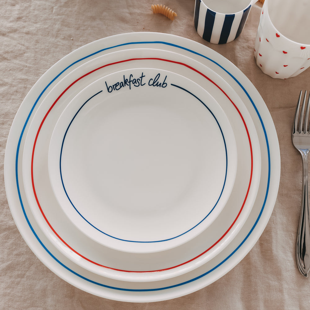 Weißer „breakfast club“-Teller mit dunkelblauem Schriftzug und farbigen Ringen, gestapelt mit weiteren Tellern auf einem beigen Leinentisch, daneben Besteck und Tassen.