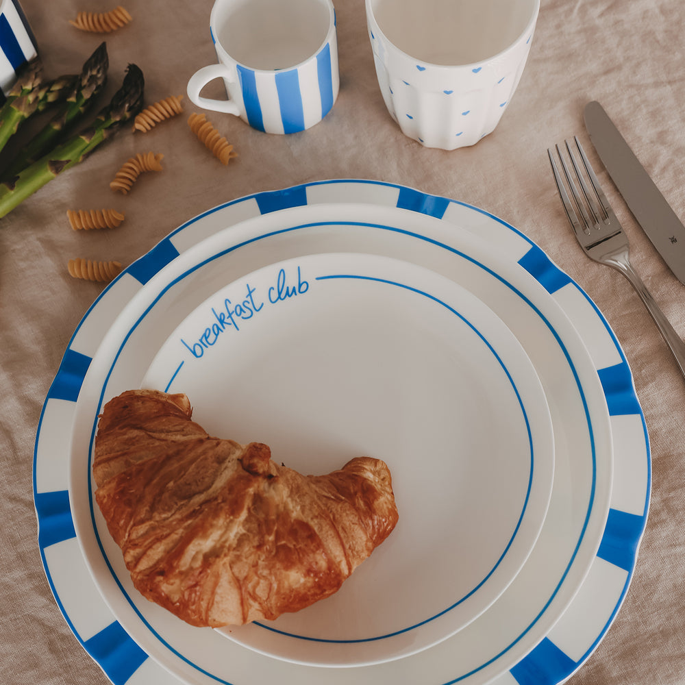 Croissant auf weißem „breakfast club“-Teller mit blauem Rand, umgeben von blau-weiß gestreiften Tassen, Tellern, Besteck und verstreuten Nudeln sowie grünem Spargel auf Leinentisch.