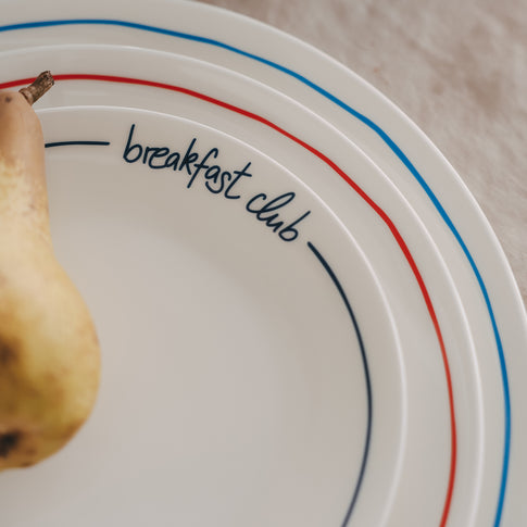 Frühstücksteller mit „breakfast club“ Schriftzug und farbigem Rand im Detail, kombiniert mit frischer Birne