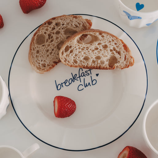 Frühstücksteller mit „breakfast club“ Schriftzug und feinem Rand in Dunkelblau von oben, dekoriert mit knusprigem Brot und frischen Erdbeeren.