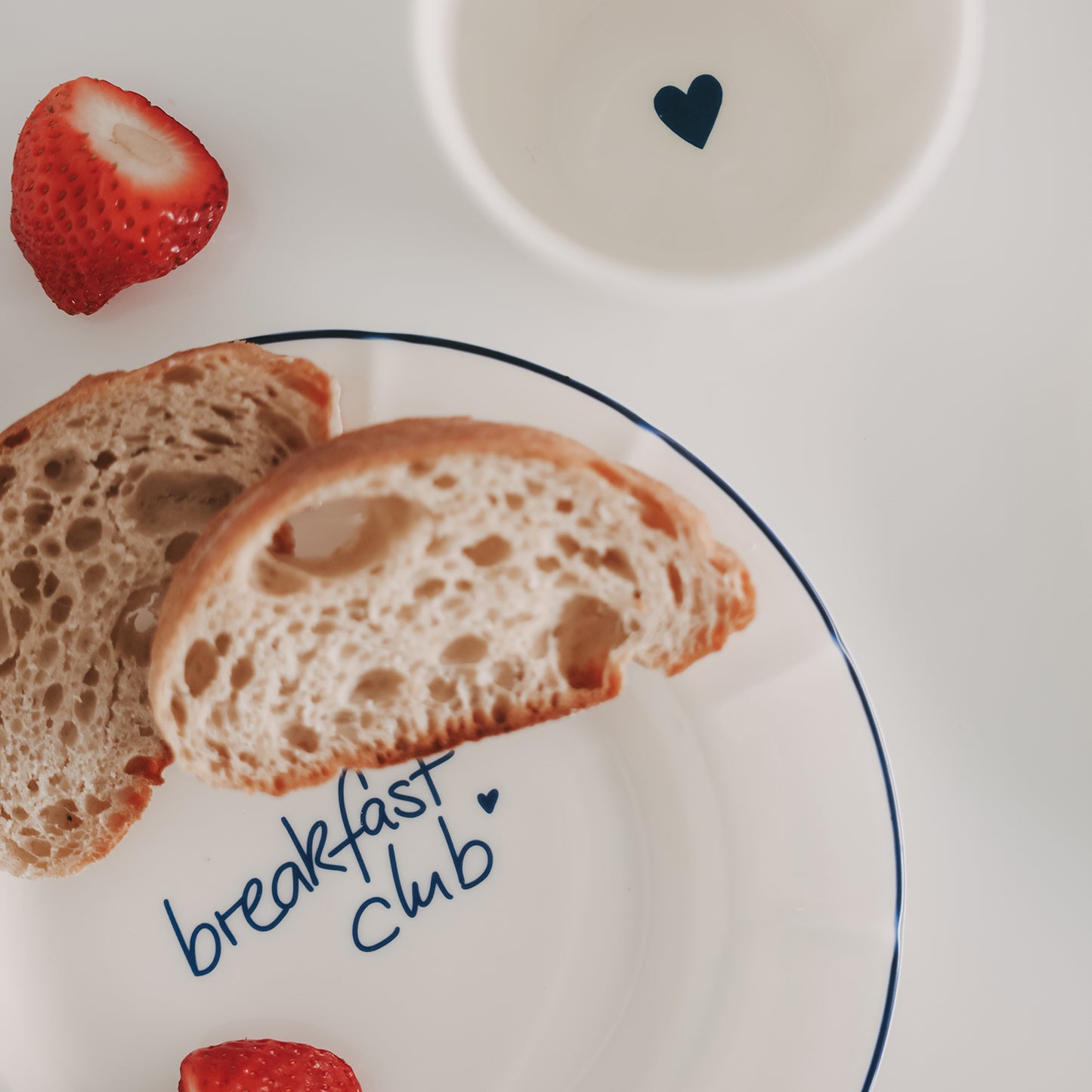Weißer Teller mit blauem Rand und Schriftzug „breakfast club“, darauf liegen zwei Brotscheiben, daneben Erdbeeren und ein Becher mit Herzmotiv.