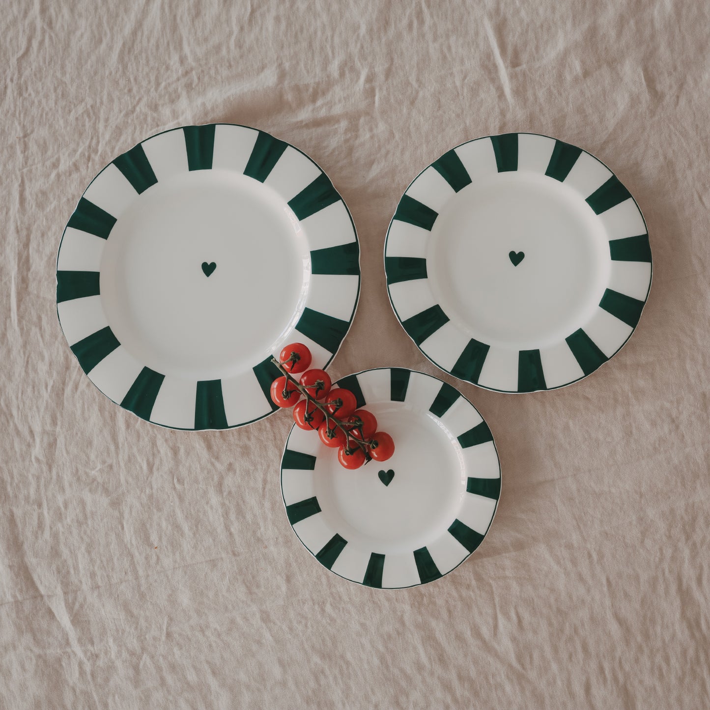 3 Porzellanteller in verschiedenen Größen mit gewelltem Rand, grünem Streifenmuster und Herzdetail, dekoriert mit frischen Tomaten auf Leinentischdecke.