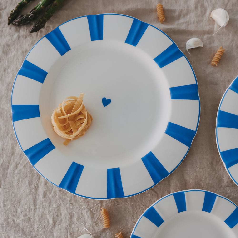 Platzteller aus Porzellan mit gewelltem Rand, hellblauen Streifen und Herzdetail, serviert mit Pasta-Nest auf Leinentischdecke