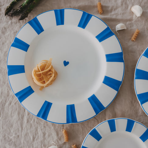 Platzteller aus Porzellan mit gewelltem Rand, hellblauen Streifen und Herzdetail, serviert mit Pasta-Nest auf Leinentischdecke