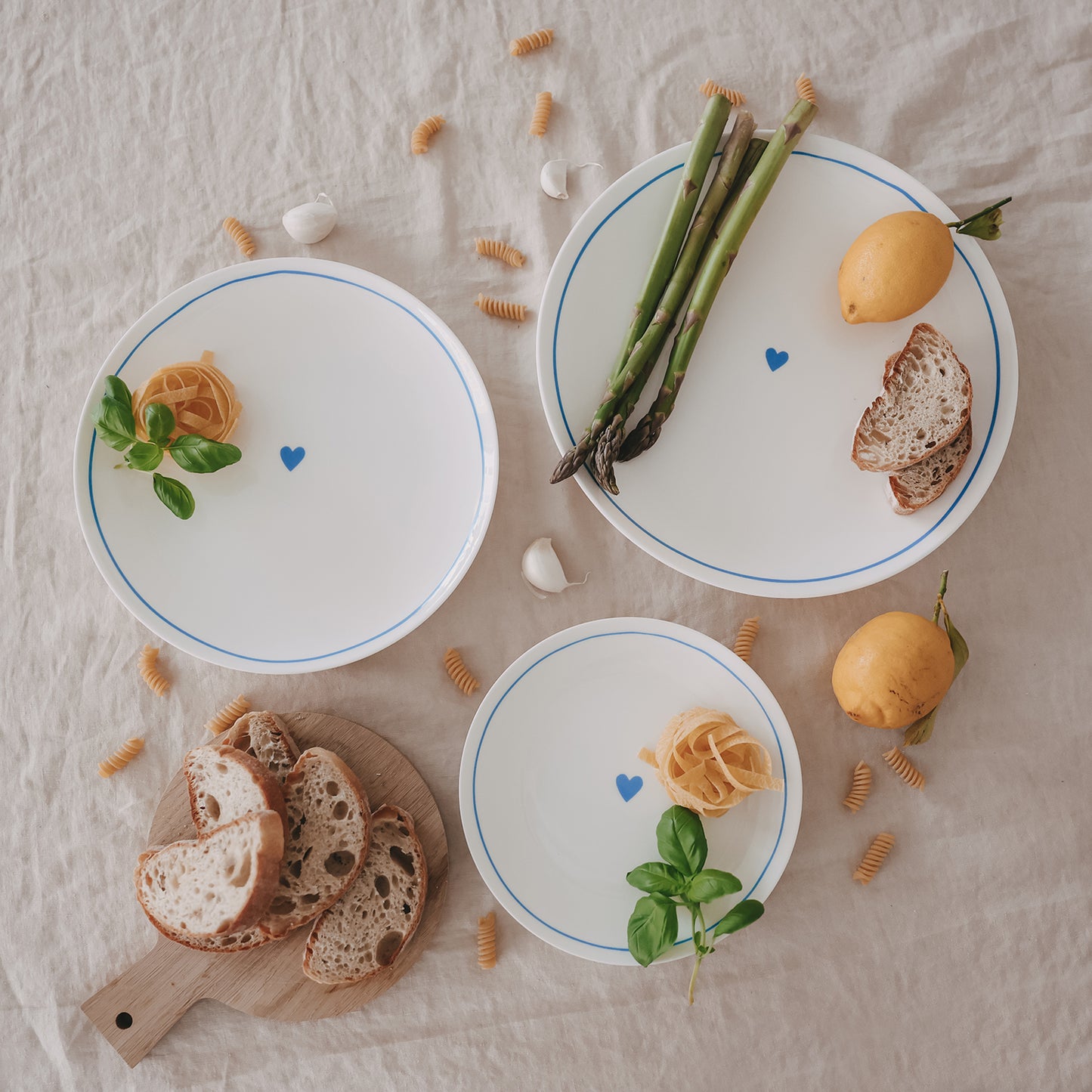 Mehrere Porzellanteller mit hellblauem Rand und Herzmotiv in verschiedenen Größen, arrangiert mit Brot, Spargel und Zitronen.